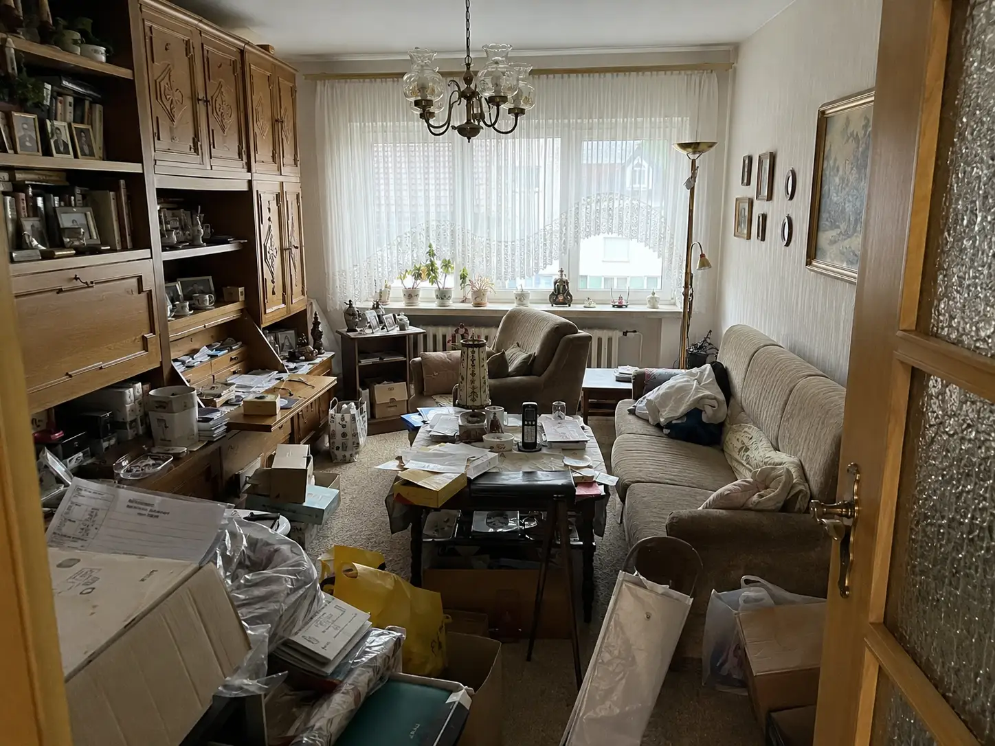 Vollgestelltes Wohnzimmer mit Schrankwand, Sofa und gestapelten Kartons und Papieren